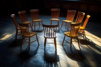 Wooden chairs arranged in circle on dark floor with dramatic sunlight casting long shadows, creating atmospheric mood for group therapy or meeting space.