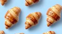 Fresh baked golden croissants arranged in rows on light blue background, top view. Flaky French pastries with glossy surface showing classic layered texture.