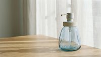 Glass soap dispenser with brass pump on wooden surface near window with natural light for bathroom or kitchen hygiene and home decor.