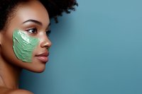 Young African American woman with natural curly hair wearing green clay mask treatment on cheek against blue background for skincare and beauty concept.