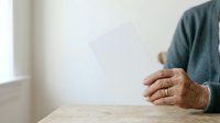 Mature man with brown skin tone holding blank white paper sheet on wooden table, suitable for mockups, presentations, and advertising campaigns.