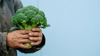 Fresh green broccoli held in weathered hands against blue background, suitable for organic food, healthy eating, and farm produce marketing materials.