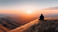 Person sitting on grassy hillside watching golden sunset over rolling hills landscape for meditation mindfulness and peaceful solitude concepts.