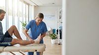 Male physiotherapist in blue scrubs treating patient leg injury in modern rehabilitation clinic with natural lighting and medical equipment.