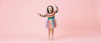 Young girl in colorful striped dress dancing with arms raised on pink background. Child expressing joy and freedom through movement and dance.