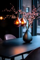 Cozy dining area with copper pendant lights and pink cherry blossoms in black vase, creating intimate atmosphere against dark modern interior design setting.