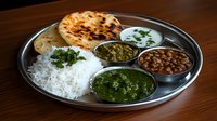 Traditional Indian thali with steamed rice, naan bread, palak paneer, chana masala, mint chutney and raita served on metal plate against wooden background.