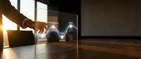 Businessman hand touching transparent digital display with glowing wave graph and data visualization in modern office workspace with wooden floor and window light.