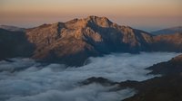 Mountain peaks emerging from sea of clouds at golden hour sunrise with warm light illuminating rocky terrain and misty valley below creating dramatic landscape.