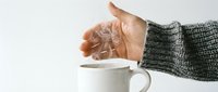 Caucasian hand in gray knit sleeve holding dandelion seedhead over white ceramic mug on white background. Concept for wishes, fragility, and natural beauty in everyday moments.