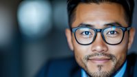 Young Asian businessman wearing glasses with confident smile in professional corporate portrait against blurred office background. Suitable for leadership concepts.