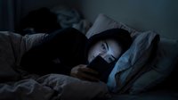 Young adult male with light skin tone lying in bed at night, illuminated by smartphone screen glow, experiencing insomnia, stress, or late-night digital addiction. Suitable for mental health, sleep disorders, technology overuse, and wellness content.