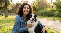 Happy young Hispanic woman with curly hair embracing her Border Collie dog in sunny park during golden hour outdoor lifestyle moment.