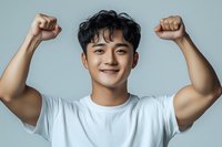 Young Asian man in white t-shirt expressing joy and triumph with raised fists against light background, showing positive emotions and success.