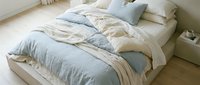 Unmade bed with blue and beige linen bedding, cream pillows, and wooden floor in bright bedroom interior for home decor and lifestyle content.