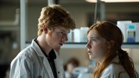 Young male with reddish-blonde curly hair and fair complexion wearing a light gray shirt converses with young female with reddish-brown hair and fair complexion in laboratory setting with blurred background and shelving with supplies.