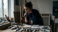 Young female artist working in creative studio space surrounded by art supplies and paintings, concentrating on artistic project with natural window lighting.
