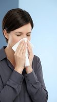 Woman with dark hair and fair complexion blowing nose with tissue while wearing dark business shirt against blue background. Suitable for healthcare, illness, allergy and cold symptom content.