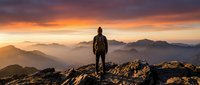 Hiker in backpack stands on rocky mountain peak at golden sunset, gazing at distant layered mountain ranges shrouded in mist and warm atmospheric light.