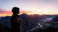 Man in brown jacket standing at railing overlooking mountain valley at sunrise, watching scenic landscape with river winding through peaks and morning mist below.