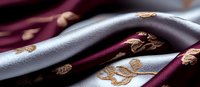 Luxurious silver and burgundy silk fabric with golden floral embroidery pattern creates elegant waves and folds in soft lighting, suitable for fashion design.