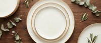Empty white plates with gold rim on wooden table with eucalyptus leaves and branches, flat lay composition for dining, tableware, and event styling projects.