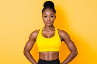 Strong African American female athlete in yellow sports bra and black leggings posing confidently against vibrant yellow background, showcasing muscular arms and fit physique.
