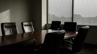 Modern corporate boardroom with dark wooden conference table and black leather executive chairs in professional office setting with window blinds.