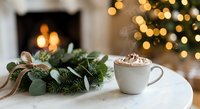 Steaming hot cocoa with whipped cream beside a festive wreath, glowing fireplace and Christmas tree in cozy winter interior.
