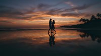 Romantic couple silhouette walking on tropical beach at sunset with dramatic sky colors reflected in wet sand and palm trees in background.