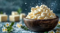 Fresh cottage cheese in rustic wooden bowl with chamomile flowers and cheese cubes on dark background, dairy product for healthy breakfast.