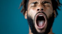 Young African American man with beard and dreadlocks yawning with eyes closed against turquoise background, expressing tiredness and exhaustion in studio lighting.