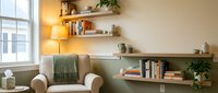 Cozy modern living room interior with beige armchair, wooden floating shelves, books, plants and warm lighting creating comfortable reading nook atmosphere.