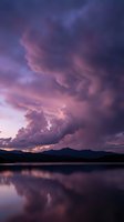 Dramatic purple and pink storm clouds reflected in calm lake water at sunset with mountain silhouettes creating moody atmospheric landscape scene.