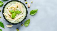 Creamy penne pasta with basil pesto sauce in dark ceramic bowl, garnished with fresh basil leaves and pistachios on light gray background. Food photography.