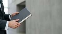 Businessman in dark suit holding closed dark book with white pages edge against blurred concrete wall background, professional education and knowledge concept for business training materials.