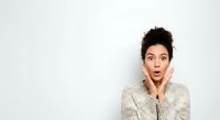 Surprised young woman with dark curly hair and medium skin tone wearing beige sweater, hand to face expressing shock or amazement against white background for advertising and marketing.