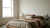 Minimalist bedroom interior with natural light from window, neutral bedding in beige and rust tones, wooden platform bed, table lamp, and potted plant for home decor and design inspiration.