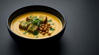 Creamy chickpea curry soup with herb-crusted tofu cubes in black bowl on dark background. Healthy vegan protein meal with fresh cilantro garnish.