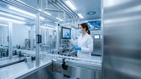 Female scientist in white lab coat working with advanced pharmaceutical manufacturing equipment in sterile cleanroom facility for drug production.