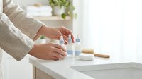 Woman in linen shirt using natural skincare products on white bathroom counter with wooden brush for daily beauty routine and self-care ritual.