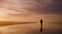Silhouetted surfer holding board standing on wet beach sand at golden hour sunset, reflecting in water for travel and lifestyle content.