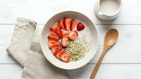 Overhead view of a white bowl containing sliced strawberries, hemp seeds, and yogurt on a wooden table with a wooden spoon and linen napkin, perfect for food blogs and healthy eating content.