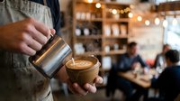 Barista pouring steamed milk into a latte art cup in a cozy coffee shop, with warm bokeh lights and customers in the background.