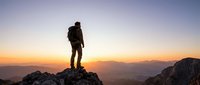 Male hiker with backpack standing on mountain peak at sunset, overlooking vast valley landscape. Adventure and outdoor exploration concept.