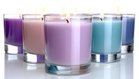 Colorful aromatic candles in glass holders arranged in row showing gradient from purple to blue tones, lit wicks glowing against white background.