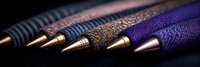 Luxury fountain pen tips in gold and copper tones against dark textured fabric background, arranged in diagonal line showing metallic nib details.