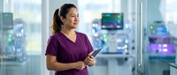 Female healthcare professional in purple scrubs holding tablet in modern hospital corridor with blurred medical monitors and equipment in background, suitable for medical, healthcare, and clinical applications.