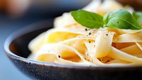 Fresh fettuccine pasta with black pepper and basil leaves served in dark ceramic plate, macro food photography with selective focus and soft lighting.