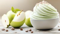 Creamy whipped dessert topping in white bowl with fresh sliced green apples and chocolate chips scattered on light surface, soft focus food photography.